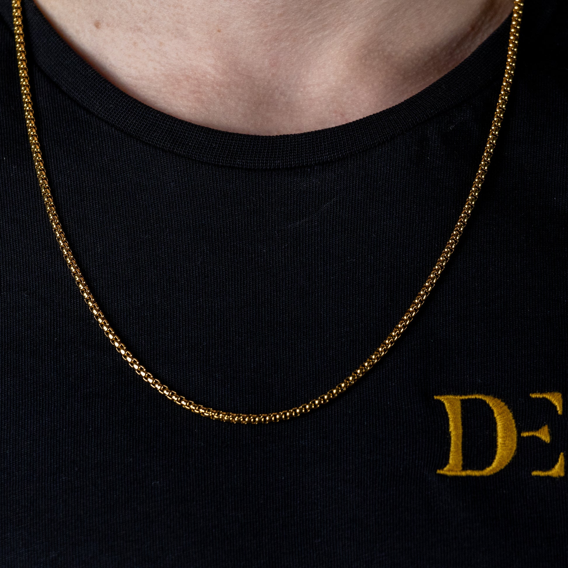 The image captures a close-up of a slender, gold chain necklace worn by someone in a black crew neck shirt with the golden lettering 'DE' visible, suggesting it may be branded apparel. The necklace's simplicity complements the implied innovative and stylish design ethos of DriftElement, a youthful German startup known for creating watches with unique rim designs, reflecting their commitment to both elegance and automotive-inspired aesthetics.